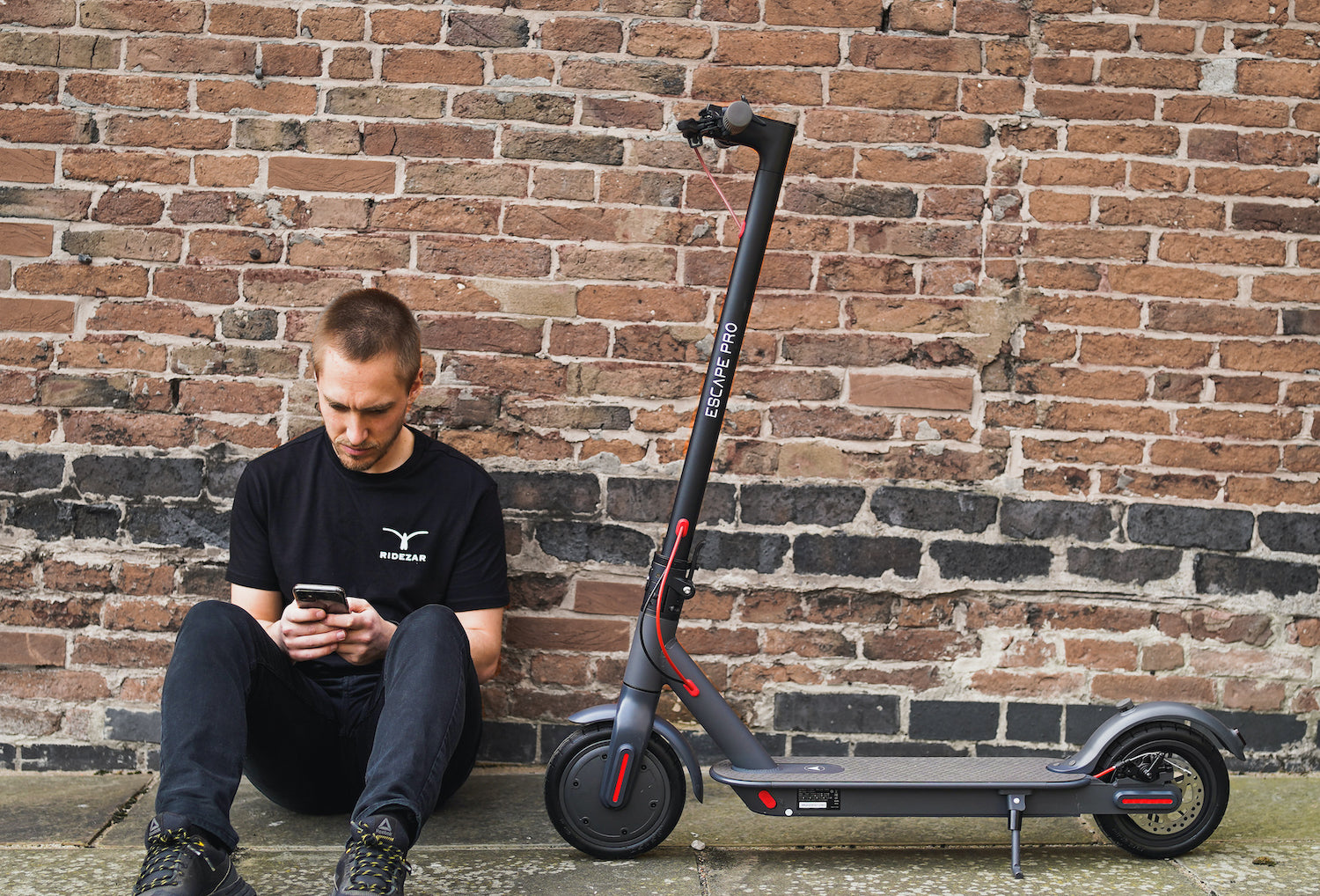 Sitting man with 30km/h Escape Pro Electric Scooter in a Ridezar T-shirt.